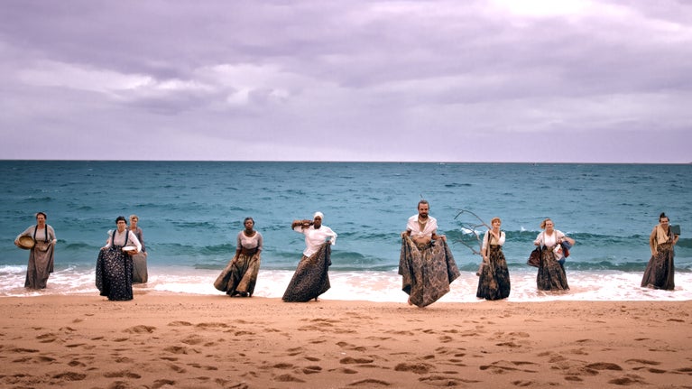 Nine performers in long skirts walking out of the sea onto a sandy beach